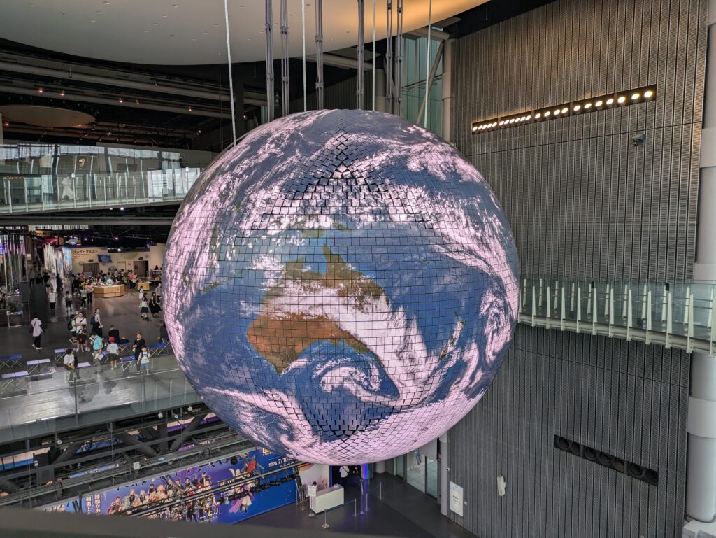 A large, glowing high-resolution LED globe hanging in the open atrium of the Miraikan museum, displaying a realistic satellite view of Earth’s clouds and oceans. A great stop on our Odaiba Day Trip. 