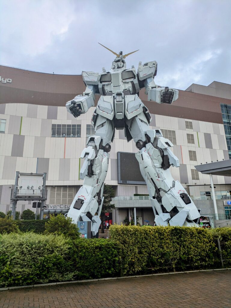 A towering, life-sized white and red Unicorn Gundam robot statue standing in front of the modern DiverCity Tokyo Plaza shopping center during out Odaiba Day Trip