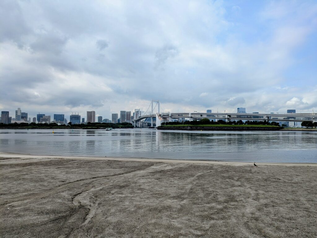 A wide-angle view of the artificial beach at Odaiba Marine Park on a cloudy day, looking out over the calm water of Tokyo Bay toward the white suspension cables of the Rainbow Bridge.