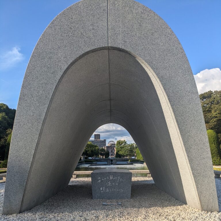 The concrete saddle-shaped Memorial Cenotaph at Hiroshima Peace Memorial Park, framing the A-Bomb Dome in the distance under a blue sky.