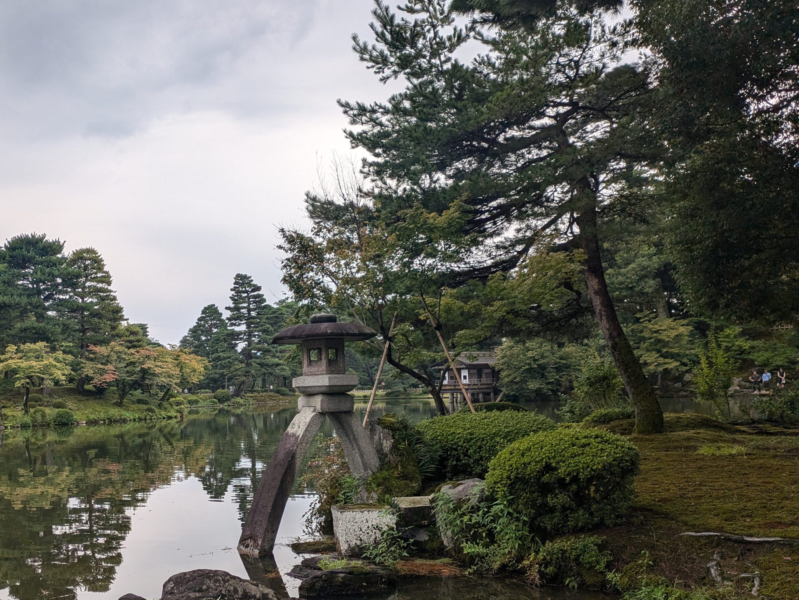 A traditional Japanese stone lantern standing at the edge of a calm pond in Kenrokuen Garden, Kanazawa, with lush pine trees and a wooden tea house reflecting in the still water.