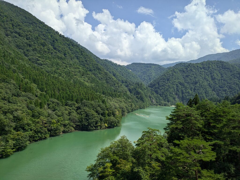 Emerald green water of a lake winding through lush, forest-covered mountains in the Japanese Alps under a blue sky with white clouds.
