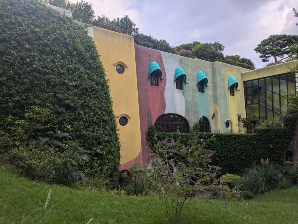 A whimsical, multi-colored building with rounded edges and bright yellow and red walls, almost entirely covered in thick green climbing ivy and surrounded by tall forest trees.