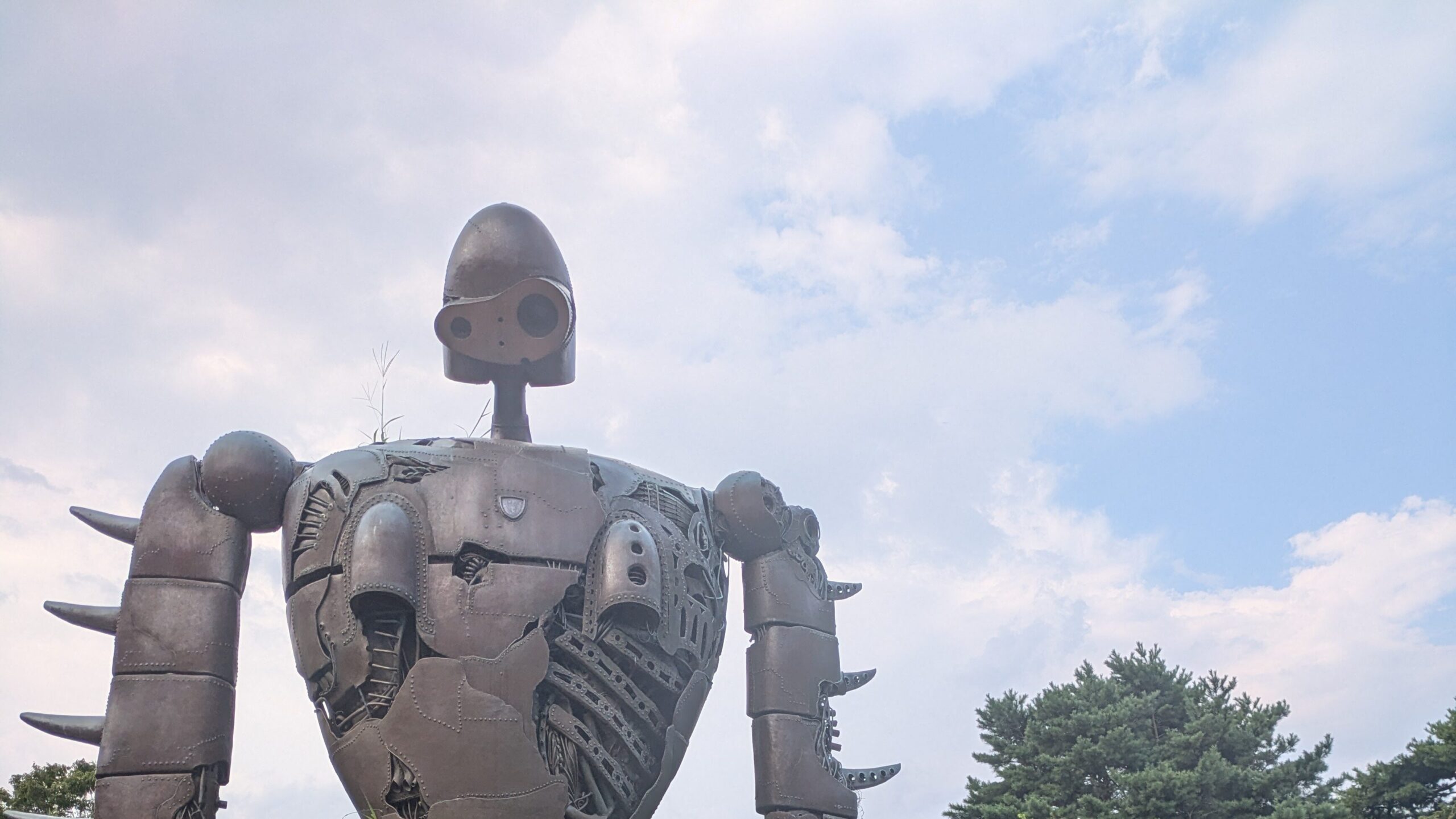 A tall, rusted bronze statue of a Laputan Robot Soldier standing amongst green bushes and grass on a rooftop, with a clear blue sky in the background.
