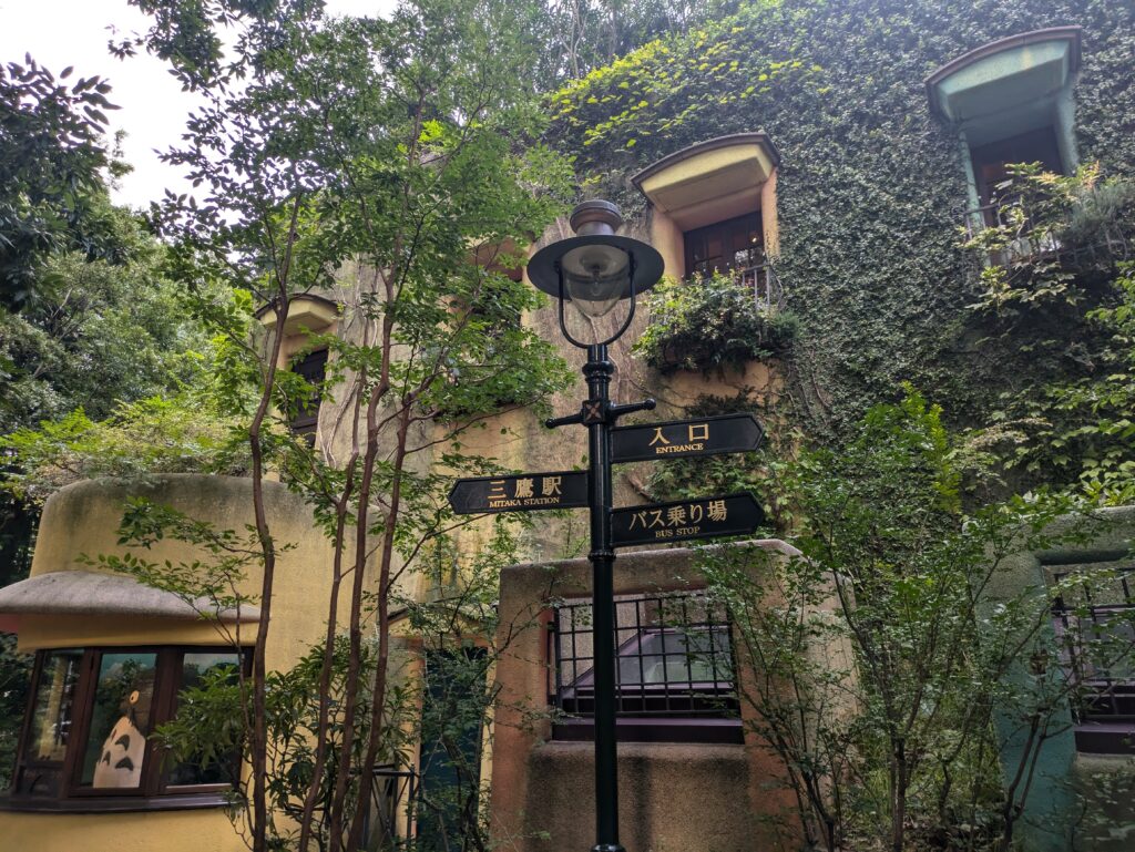 A black directional signpost in front of the Ghibli Museum's yellow, ivy-covered walls. To the left, a large Totoro figure is visible inside a circular window.
