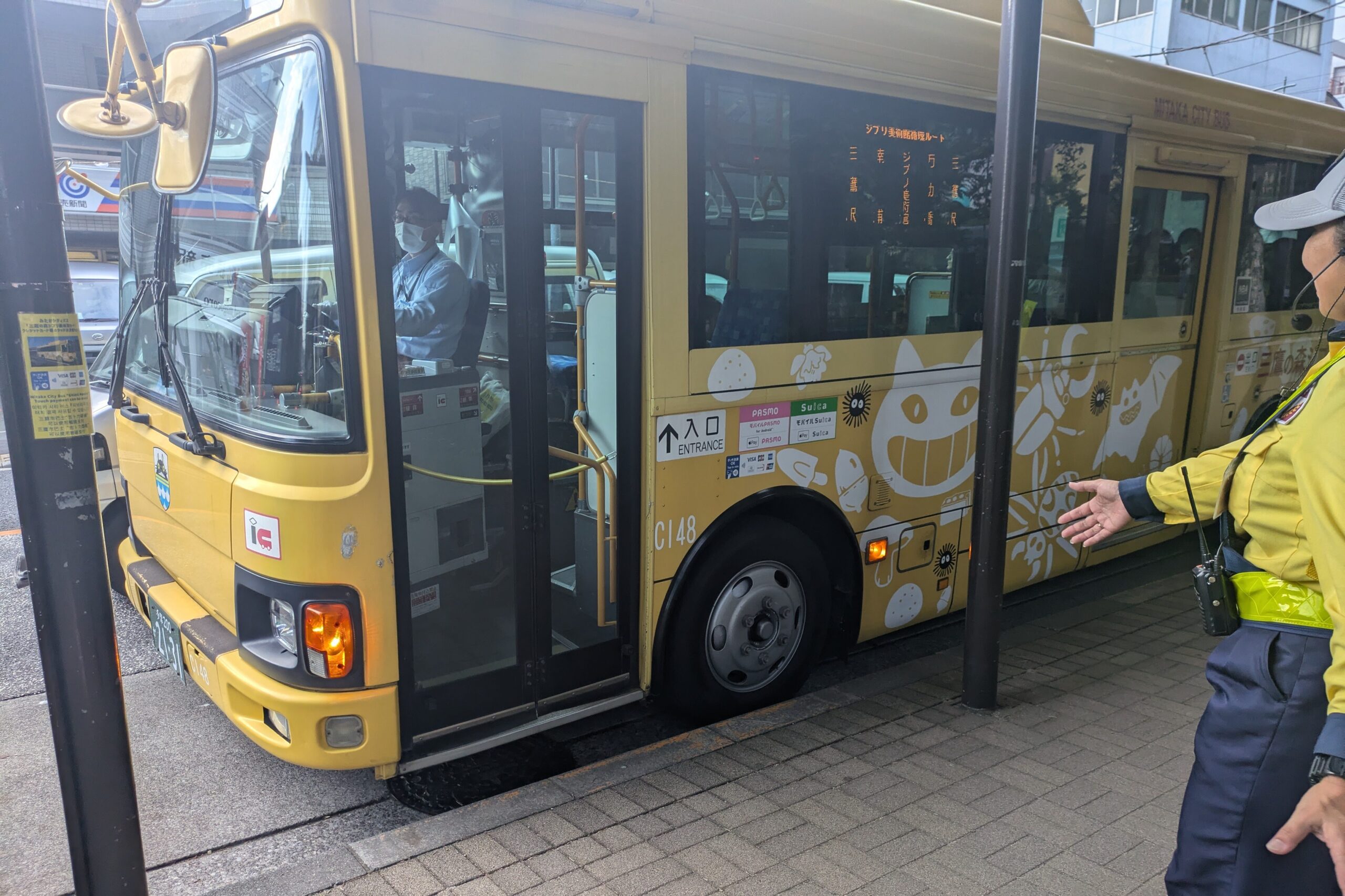 A bright yellow public shuttle bus at Mitaka Station in Japan, featuring white illustrations of Studio Ghibli characters like Catbus and soot sprites on its side.