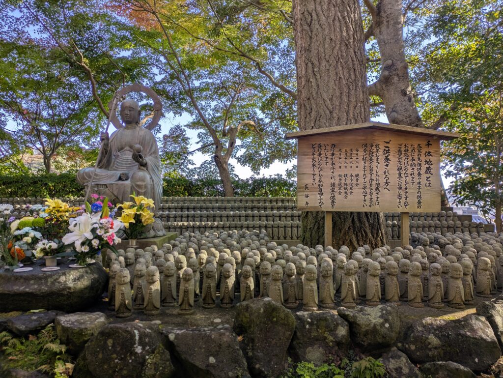 A close-up of hundreds of small, stone Jizo statues with peaceful expressions, some covered in green moss, huddled together on a stone ledge in a garden.