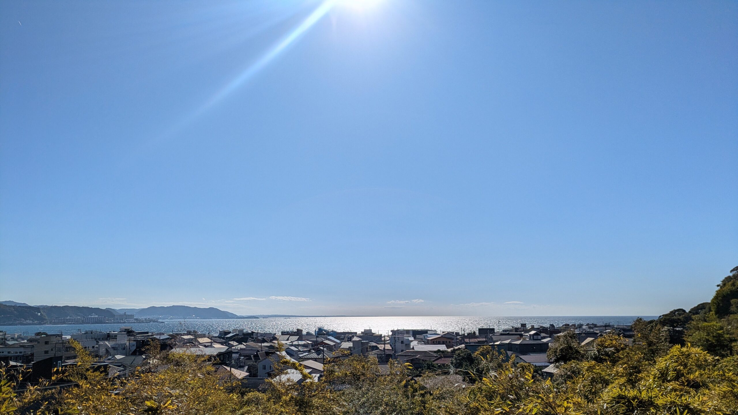 A high-angle panoramic view from a wooden temple balcony at Hasedera, looking out over a coastal Japanese town and the blue crescent-shaped bay under a bright sky.