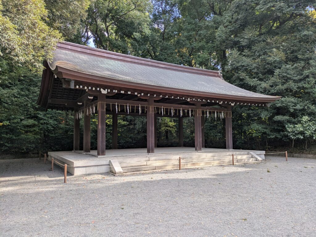 A traditional Japanese wooden shrine pavilion nestled deep within a forest of tall, straight cedar trees, highlighting the architectural symmetry and serene natural setting.