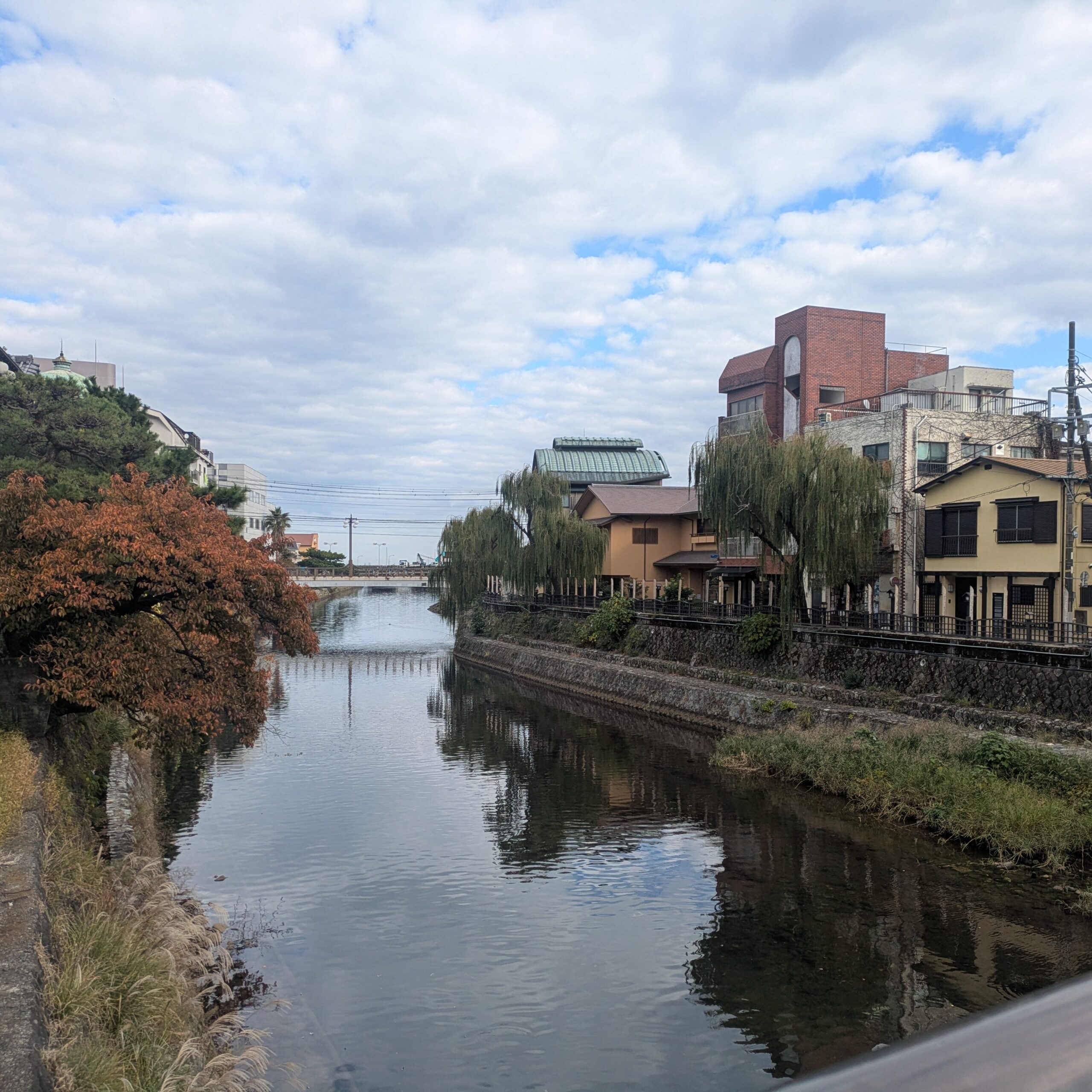 A scenic view of the Matsukawa River in Ito with autumn trees, traditional buildings, and weeping willows reflecting in the calm water.
