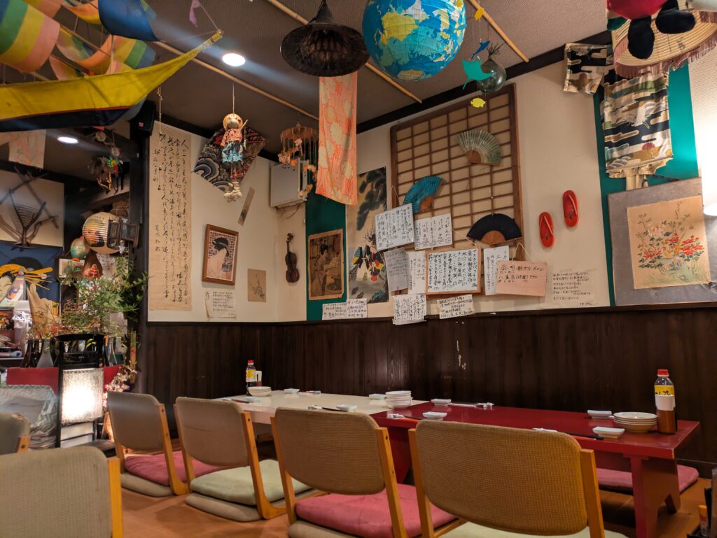 The eclectic interior of Kunihachi Izakaya in Ito, featuring low floor seating with cushions, wood-paneled walls covered in handwritten notes and drawings, and various traditional Japanese knick-knacks hanging from the ceiling.