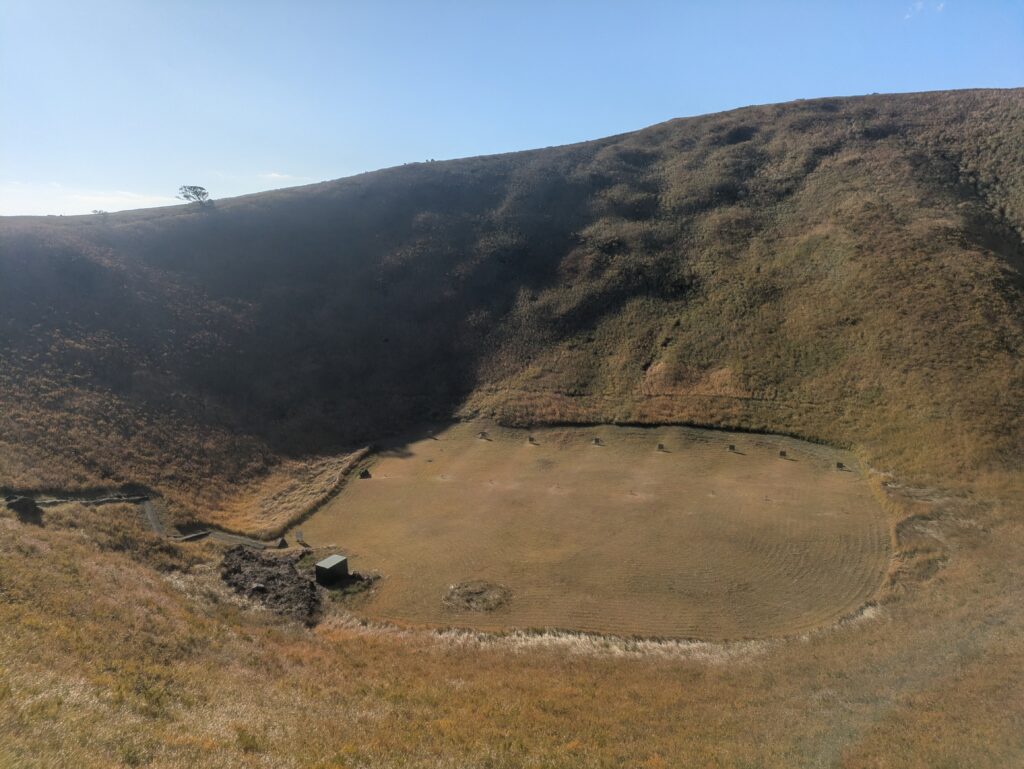 A wide, panoramic view of the deep, grass-covered crater at the summit of Mt Omuro in Izu, Japan, showing the walking path along the rim.