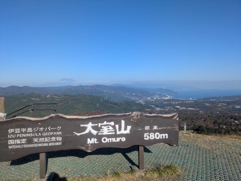 A scenic view of the snow-capped Mt Fuji in the distance, framed by the wooden Mt Omuro summit sign in the foreground on a clear, sunny day.