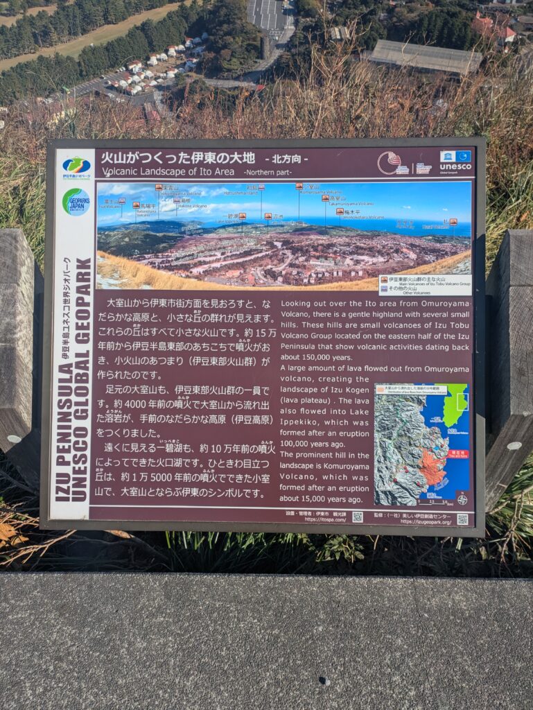 A close-up of a Japanese and English informational sign at the top of Mt Omuro explaining the Izu Peninsula UNESCO Global Geopark and its volcanic history.