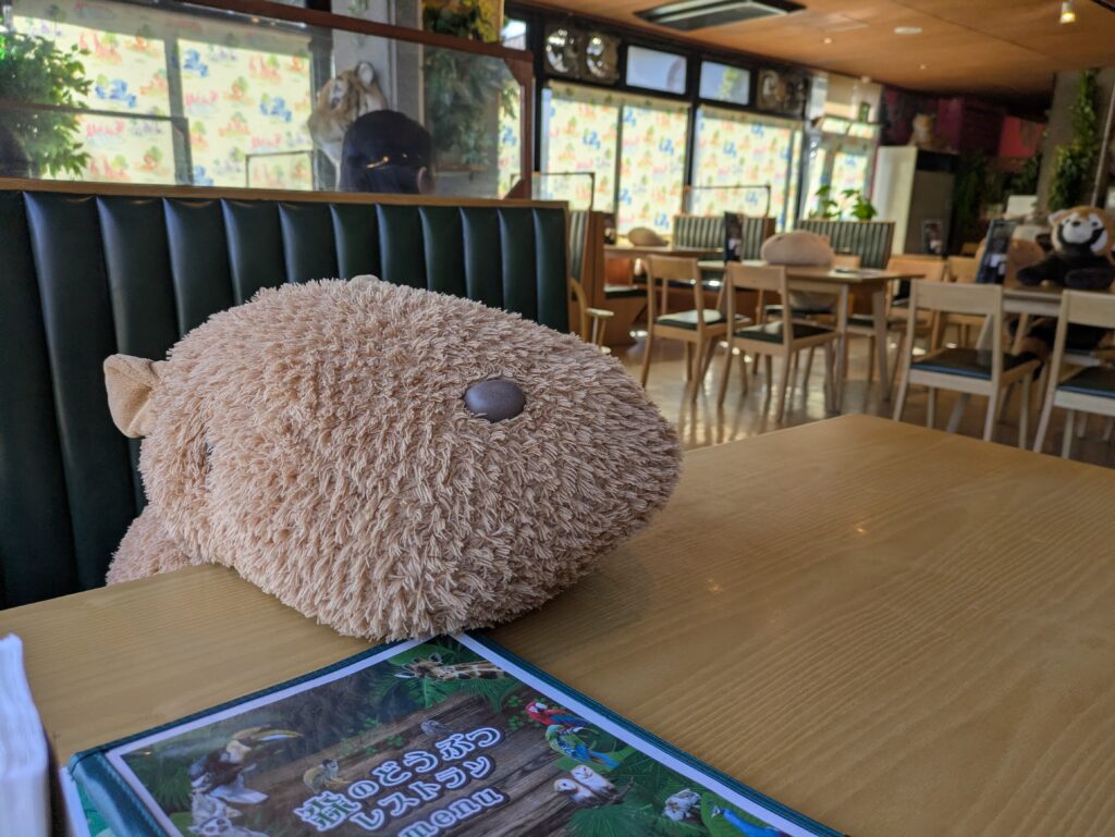A large, fuzzy brown capybara plushie sitting on a wooden table at the Izu Shaboten Zoo restaurant, with a cafe interior and empty tables in the background.