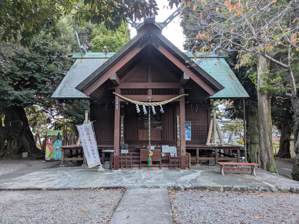 The wooden front of Otonashi Shrine in Ito, featuring a traditional straw rope (shimenawa) and paper tassels, surrounded by lush green trees.