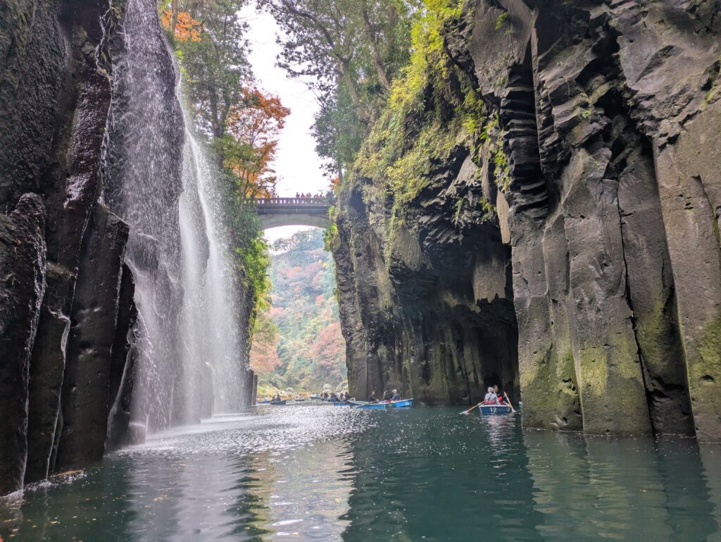 A low-angle view from a rowing boat on the water of Takachiho Gorge, looking up at a high stone bridge and the Manai Falls cascading down moss-covered basalt columns, with other small boats in the distance.