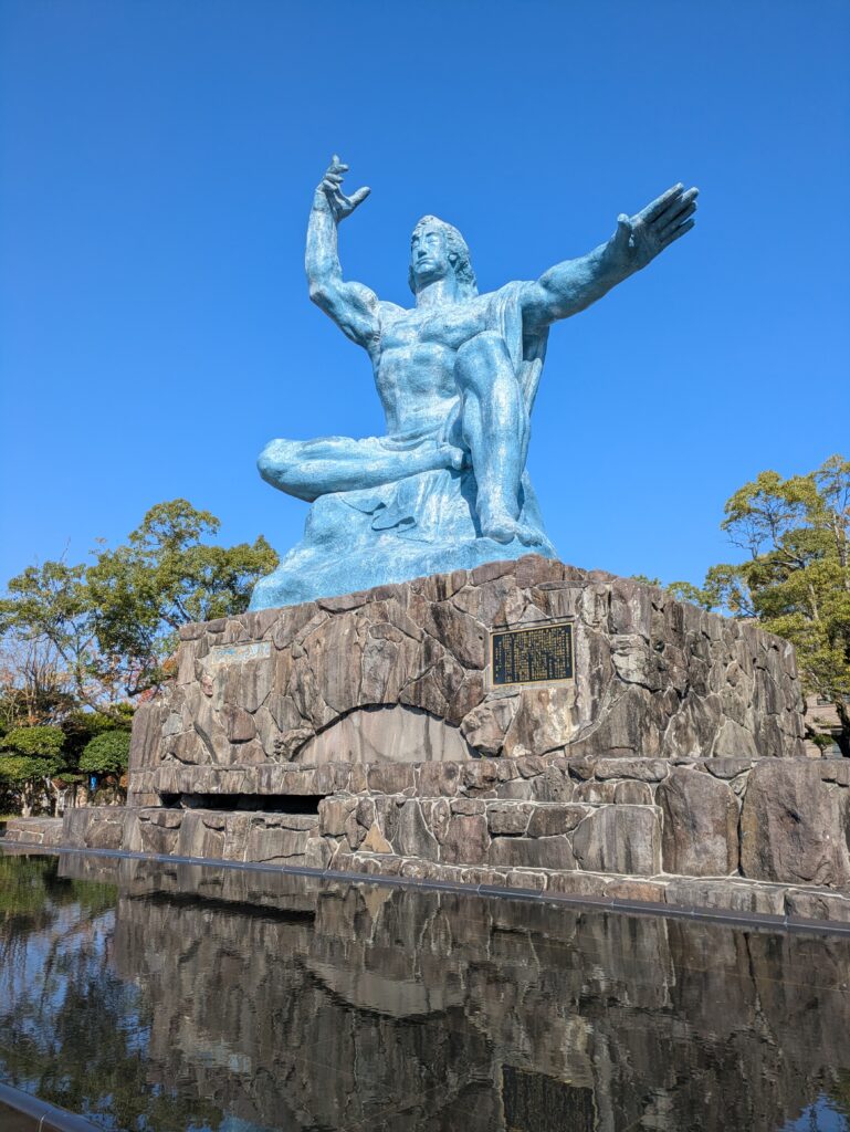 The iconic blue Peace Statue in Nagasaki Peace Park under a clear sky, reflecting in the fountain pool below.