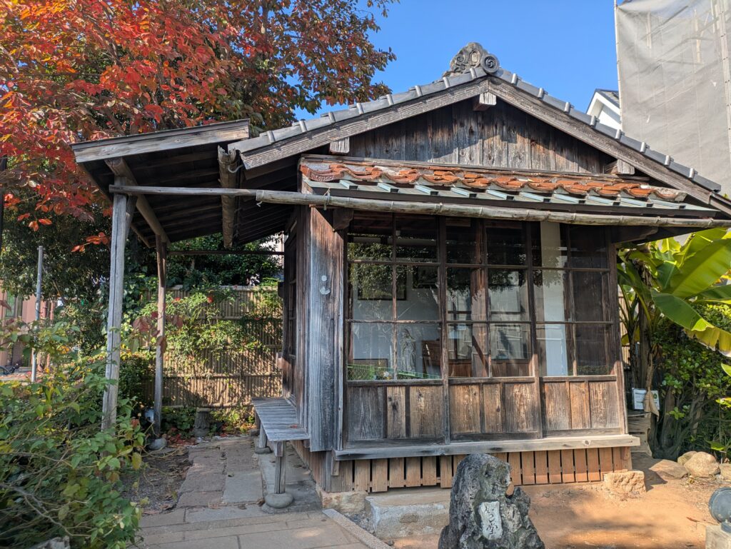 A small, humble, single-story wooden Japanese hut with traditional glass doors and sliding panels, surrounded by trees with autumn foliage.