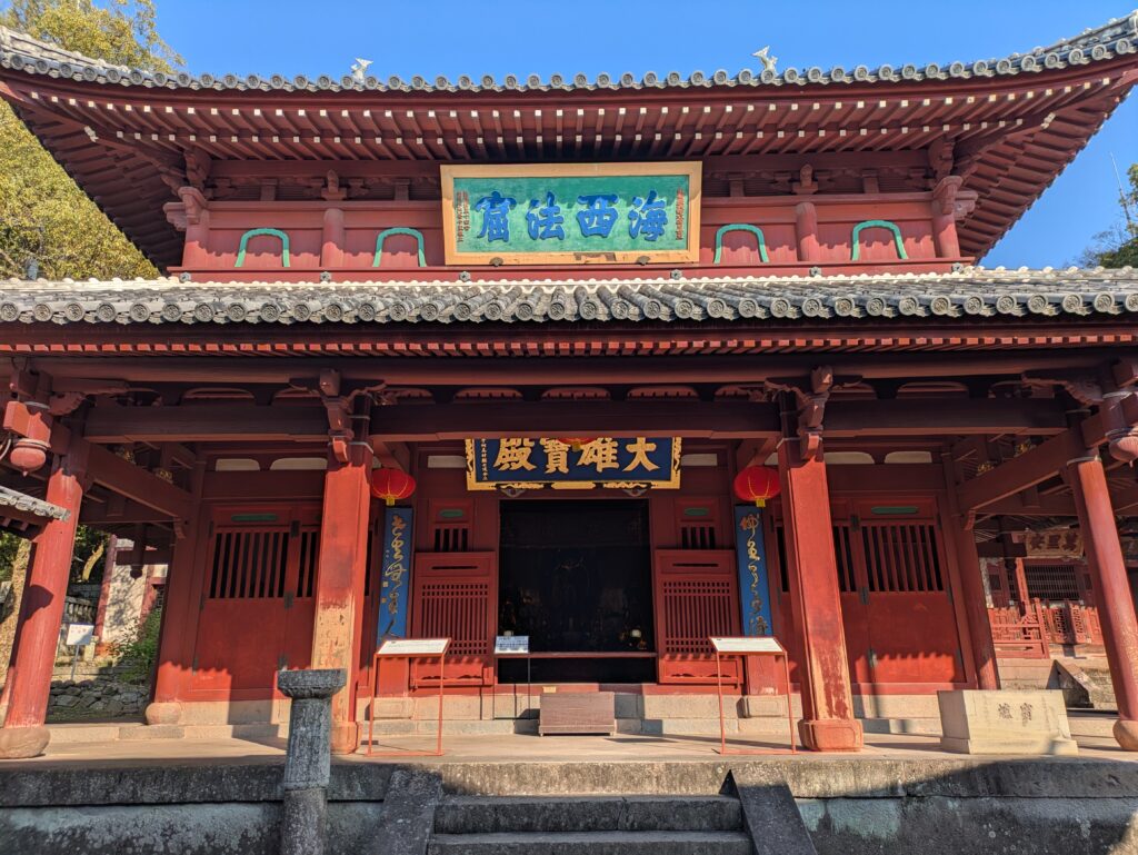 The ornate red wooden exterior of the Daiohoden (Buddha Hall) at Sofukuji Temple, featuring intricate Chinese-style carvings and a blue-and-gold framed sign above the entrance.