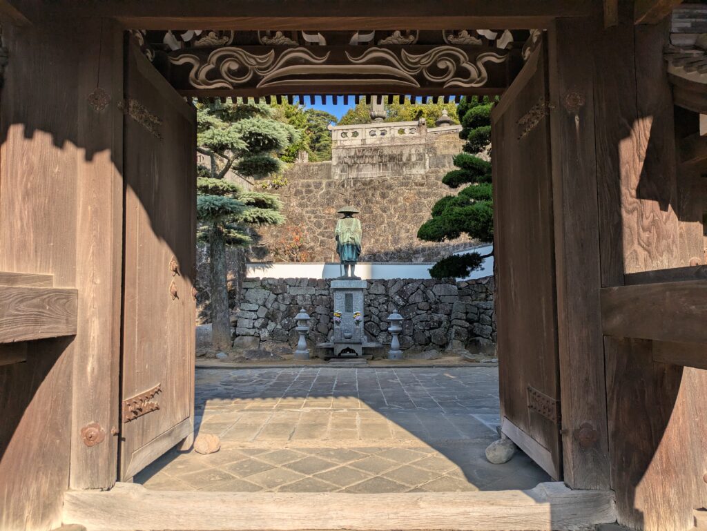 A view looking through the weathered wooden gates of Kofukuji Temple in Nagasaki, framing a stone courtyard, a central statue, and a steep hillside wall in the background.