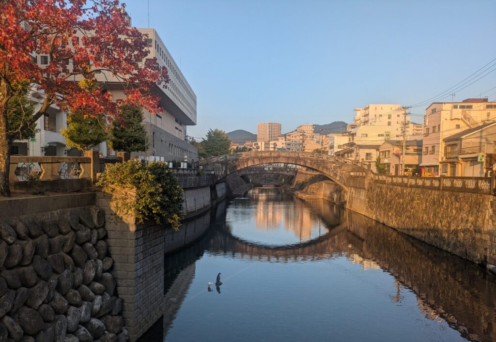 A scenic view looking down the Nakashima River in Nagasaki at an arched stone bridge, with a perfect reflection of the arches in the calm water, flanked by river walls and autumn trees under a blue sky.