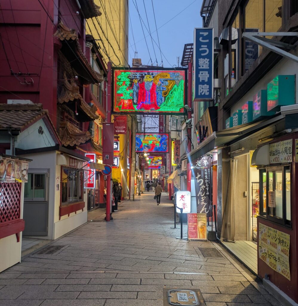 A narrow, glowing street in Nagasaki Chinatown at dusk with colorful illuminated signs and traditional architecture.