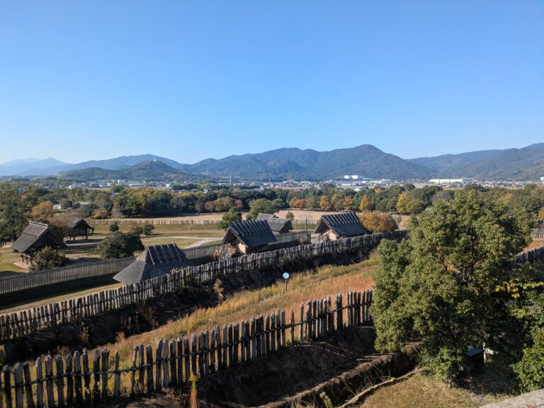 Wide panoramic view of reconstructed Yayoi period watchtowers and thatched-roof huts at Yoshinogari Historical Park with mountains in the background.