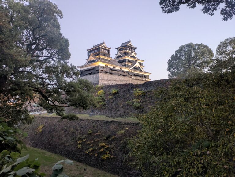 Kumamoto Castle at dusk, featuring the iconic black exterior of the main keep illuminated against a soft evening sky, framed by lush green trees and stone fortifications.