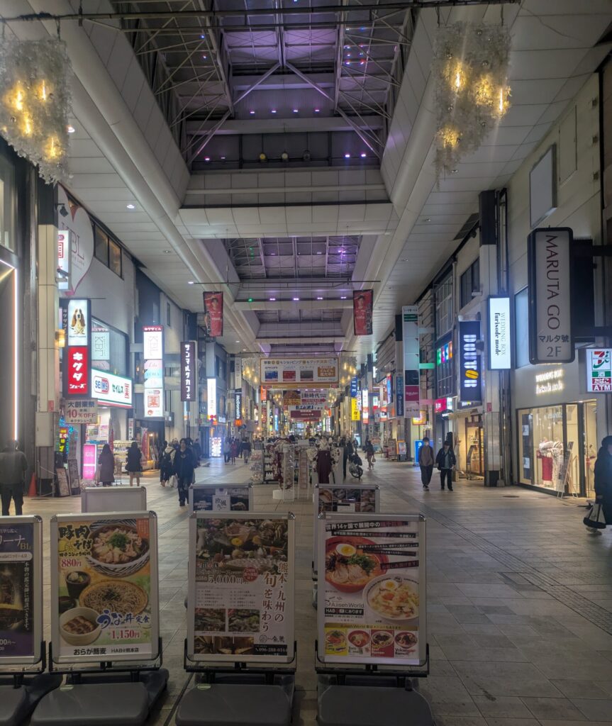 A wide-angle view of a brightly lit, covered pedestrian shopping arcade in Kumamoto, Japan. The arcade features a high, modern glass ceiling, various shop signs in Japanese and English, and street food menu boards in the foreground. People are seen walking through the spacious, clean walkway.
