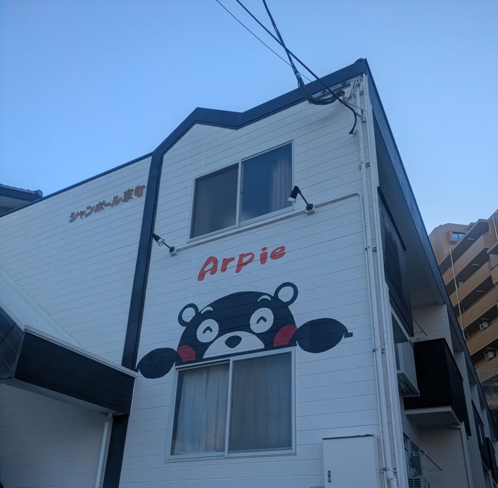 Charming view of the white facade of the Arpie accommodation in Kumamoto, featuring a friendly, large mural of the Kumamon bear peeping over a window against a clear blue sky.