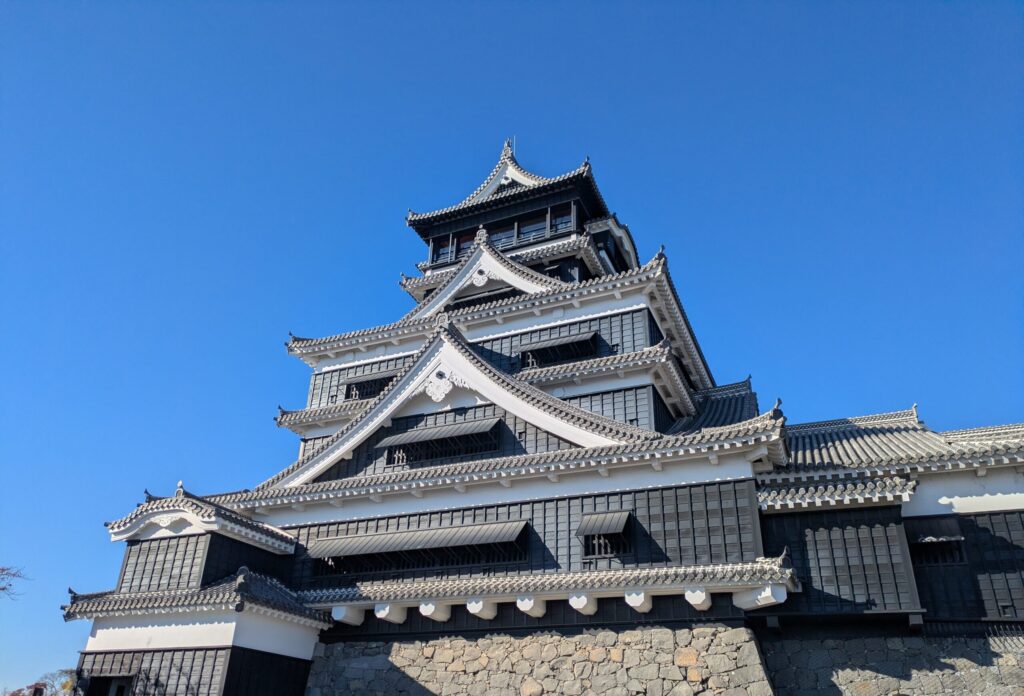 The black exterior and massive sloping stone walls of Kumamoto Castle under a clear blue sky in Kyushu, Japan.