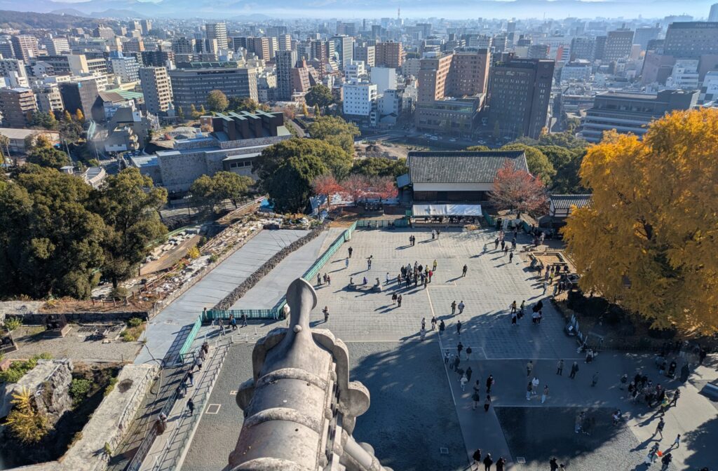 High-angle view from the top of Kumamoto Castle overlooking the main courtyard and the city skyline, featuring a large yellow ginkgo tree and traditional roof tiles in the foreground.