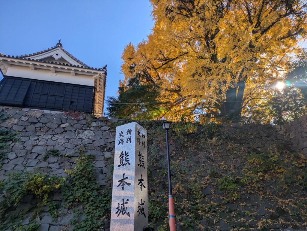 A white Japanese stone boundary marker sits on a grassy slope with stone walls. Behind it, a traditional castle turret building is partially visible. The scene is dominated by a large, massive ginkgo tree with brilliant, golden-yellow autumn leaves. A decorative lamp post is positioned next to the marker. The sky is deep blue, lit by the low sun.