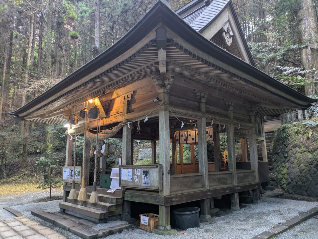 Front view of the main wooden pavilion of Kamishimi-kumano-za Shrine with illuminated lanterns at dusk.