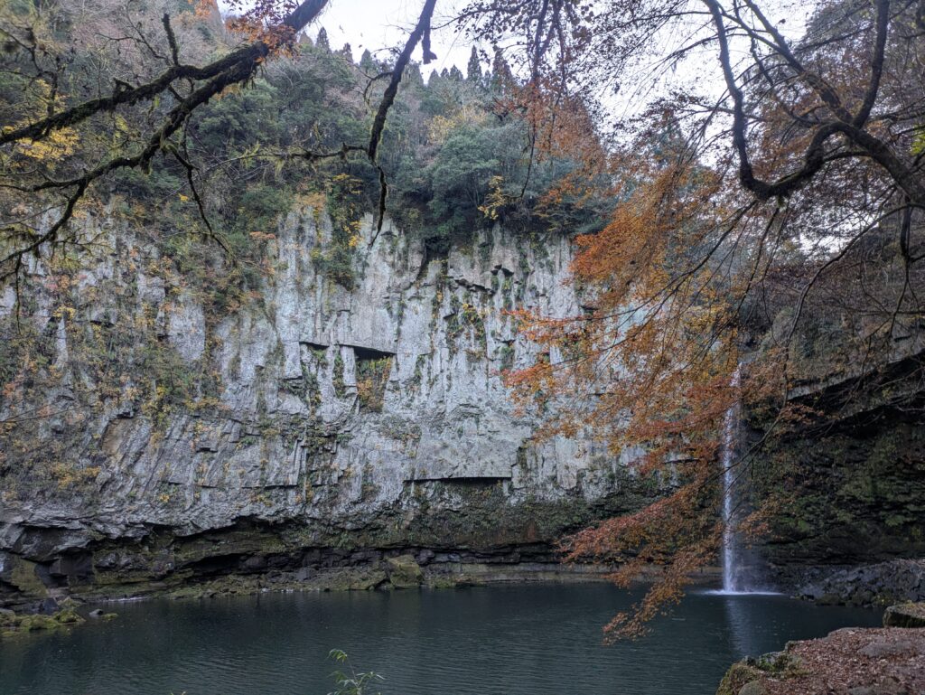 Vertical view of Unoko Falls in Gokase Japan with autumn leaves framing a high cliff face and a pond.