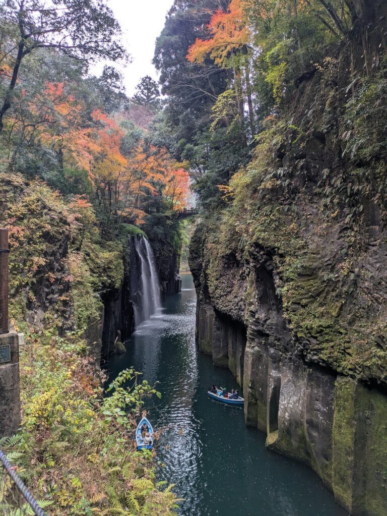 Vertical shot looking down into Takachiho Gorge, Miyazaki, Japan, showing high basalt cliffs, Manai Falls, and small rowboats on turquoise water.