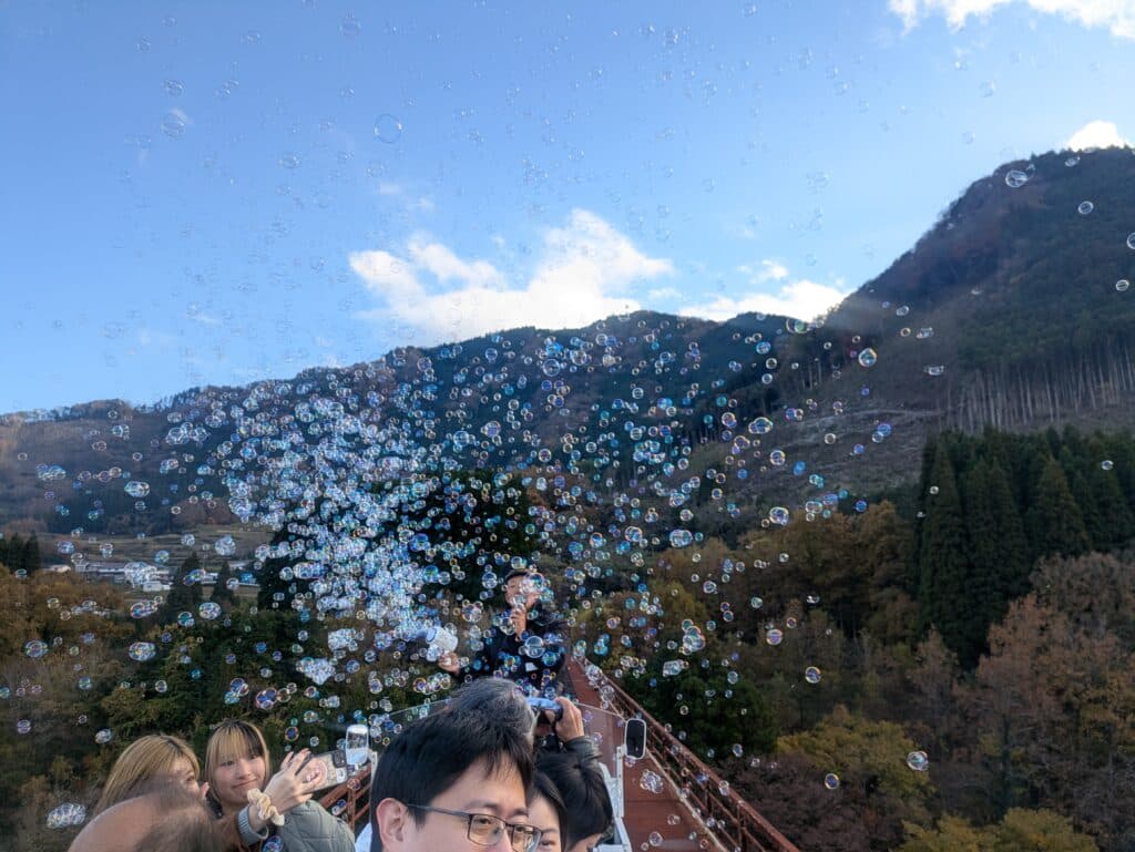 High-angle view from the Amaterasu Railway bridge overlooking a deep forested valley and a village, with white bubbles floating over the view.