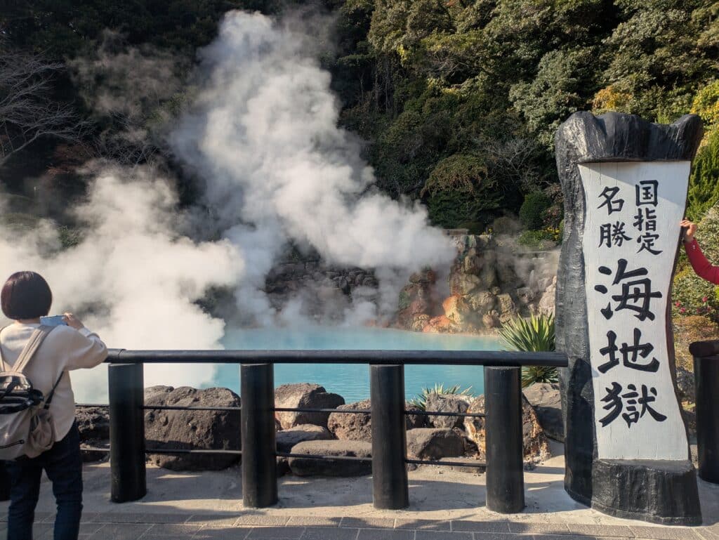 Bright turquoise hot spring pool with thick white steam rising, surrounded by dark rocks and a black wooden fence at Umi Jigoku in Beppu, Japan.