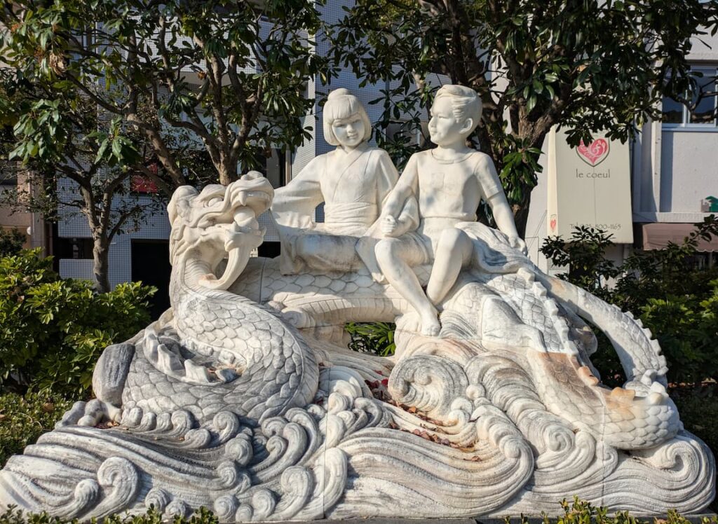 A white stone sculpture in Nagasaki of a Japanese girl and a Chinese boy sitting on the back of a large dragon leaping over waves, surrounded by green trees and a city building in the background.