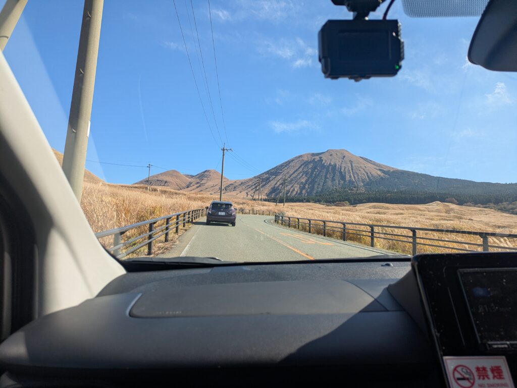 View from inside a rental car driving along a quiet, scenic road toward a volcanic mountain peak in the Mt Aso region of Kyushu, Japan.