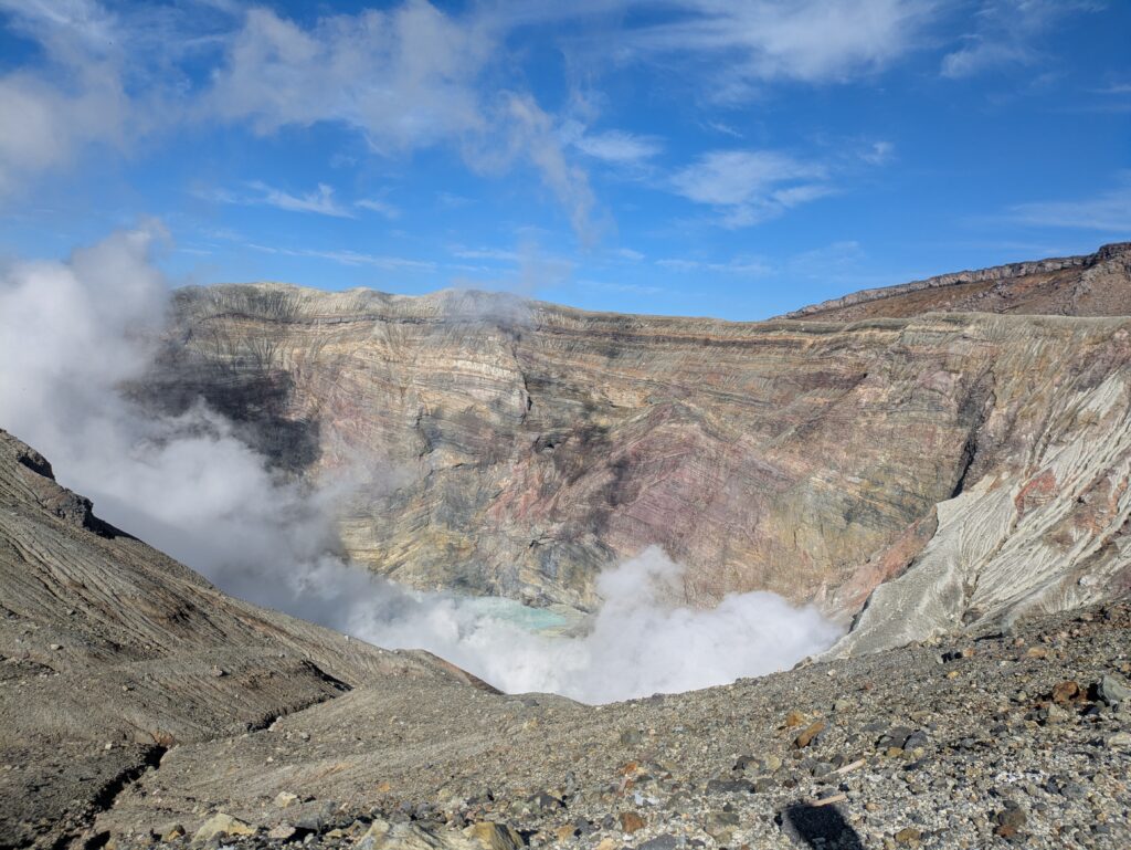 Plumes of thick white steam rising from the rugged rocky depths of the active Nakadake Crater No. 1, revealing a pale turquoise acid pool at the bottom.