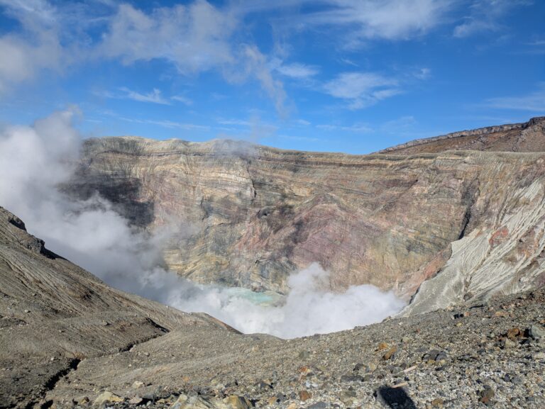 Plumes of thick white steam rising from the rugged rocky depths of the active Nakadake Crater No. 1, revealing a pale turquoise acid pool at the bottom.