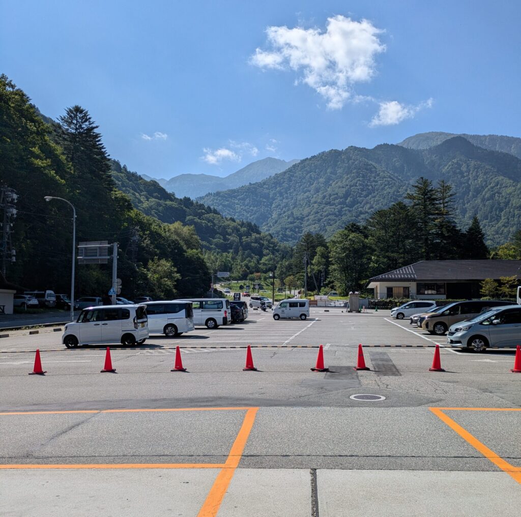 An open asphalt parking lot with orange painted lines and a row of bright red traffic cones in the foreground. Several cars and small white vans are parked beneath towering, densely forested green mountains that frame a distant mountain ridge under a clear blue sky with a few light clouds.