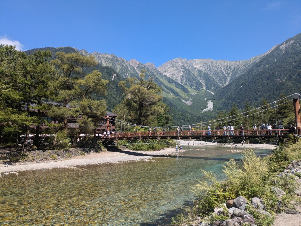 The iconic wooden Kappa Bridge spanning across the crystal-clear Azusa River in Kamikochi, Japan. People are walking across the bridge, framed by lush pine trees and the massive, rocky peaks of the Hotaka Mountain Range under a clear blue sky.