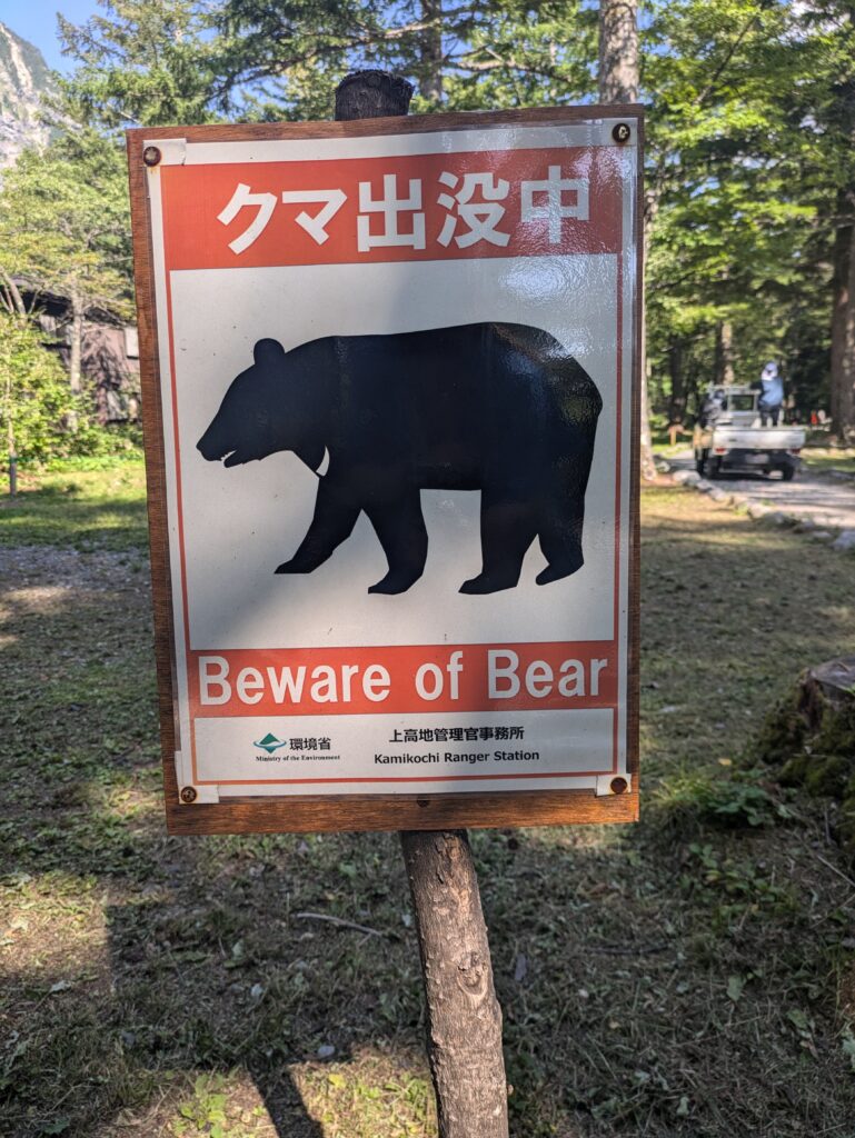 A rectangular metal sign mounted on a wooden post along a trail in Kamikochi, Japan, warning hikers about wildlife. The sign features a large black silhouette of a bear in the center, with a bright orange header containing Japanese text and a matching orange footer that reads "Beware of Bear" in English.