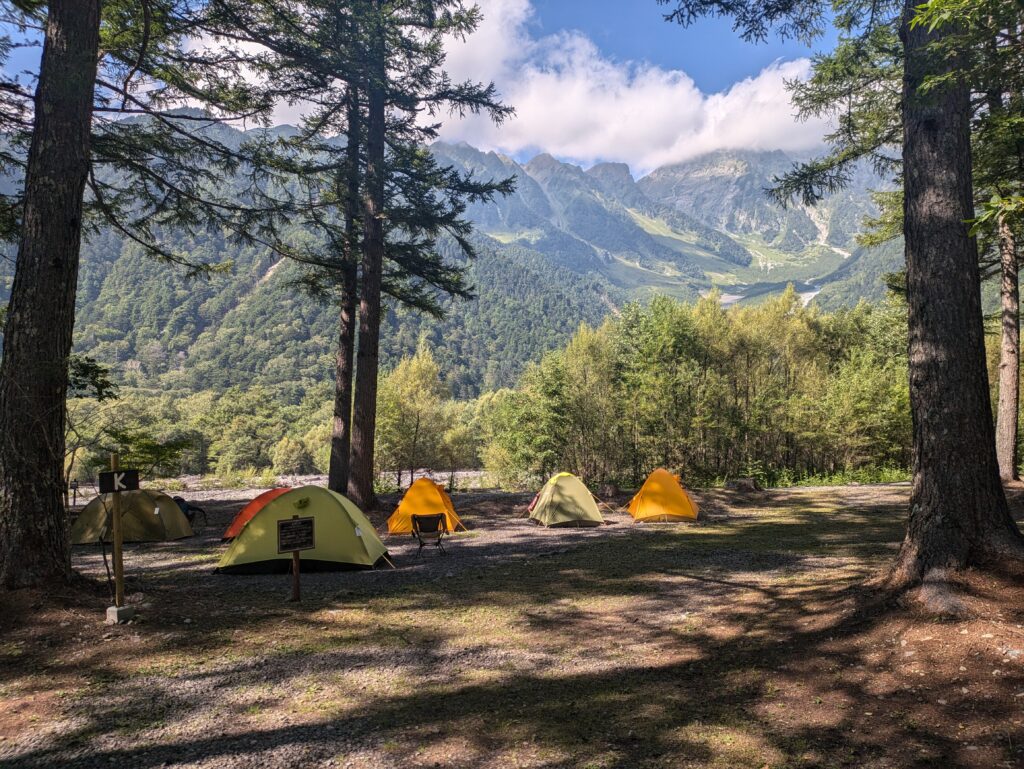 A row of small, brightly colored camping tents pitched at the Konashidaira Campsite in Kamikochi, Japan. The yellow and orange tents sit on the forest floor under tall pine trees, looking out across the riverbed toward the massive, steep peaks of the Japanese Alps under a blue sky.