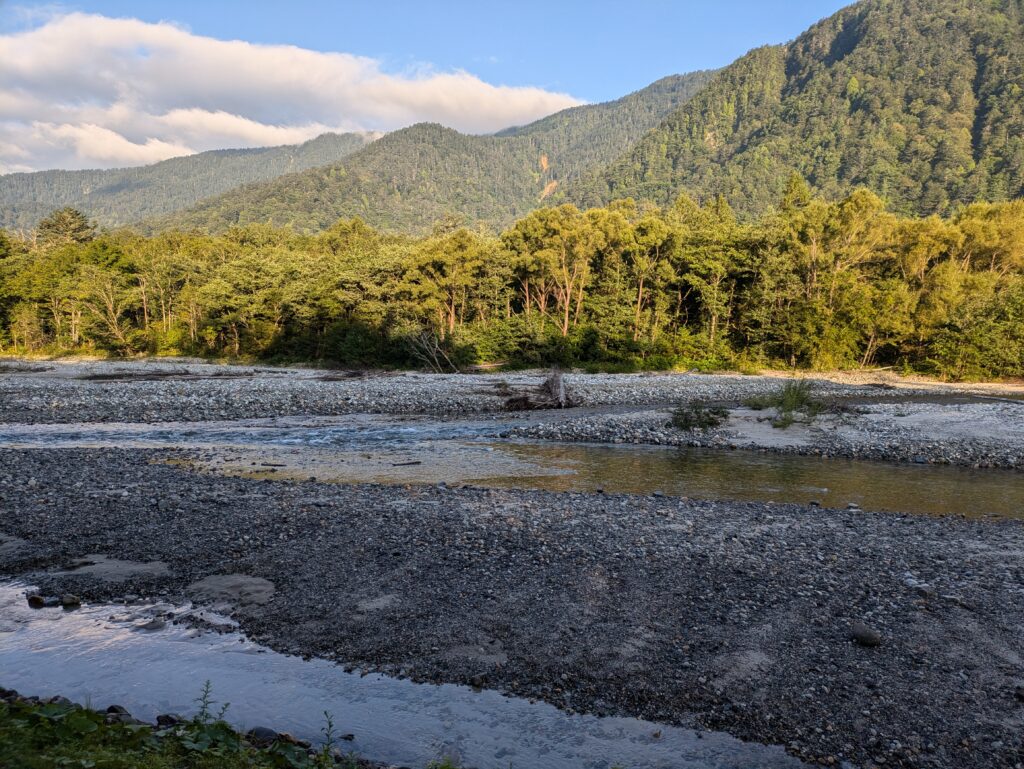 The wide, shallow Azusa River flowing over a rocky bed in Kamikochi, Japan, backed by a thick forest of green trees and rolling mountain ridges under a partly cloudy sky.