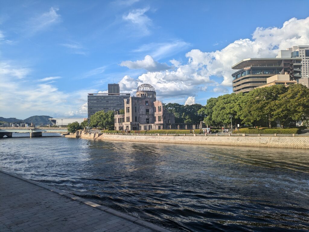 The Genbaku Dome (Atomic Bomb Dome) in Hiroshima, seen from across the river under a blue, cloudy sky.